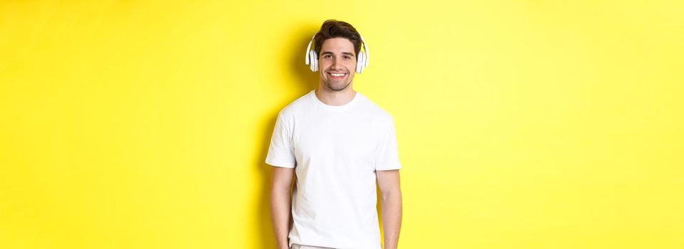 Young handsome man listening music in headphones, wearing earphones and smiling, standing over yellow background.