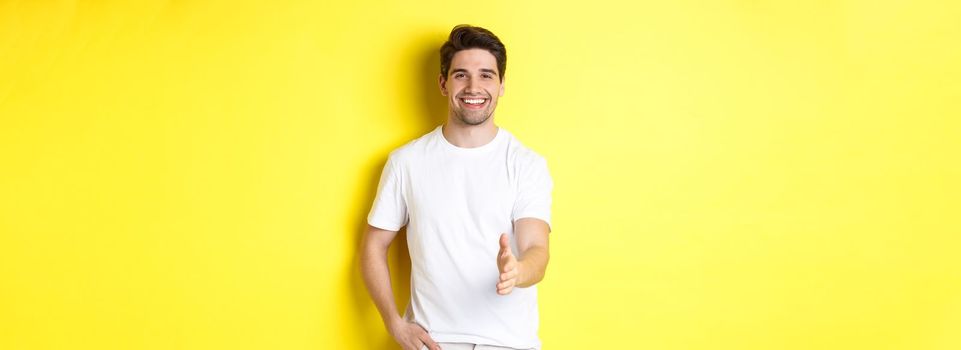 Handsome and confident man extending hand for handshake, greeting you, saying hello, standing in white t-shirt over yellow background.