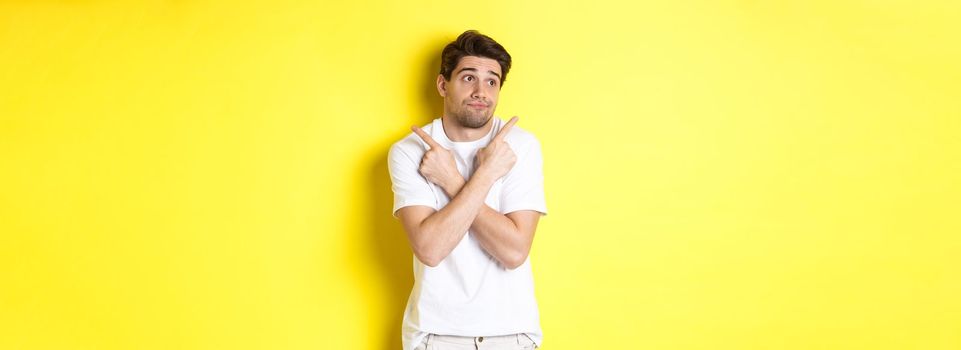 Indecisive man shrugging, pointing fingers sideways, troubled to choose, standing over yellow background.