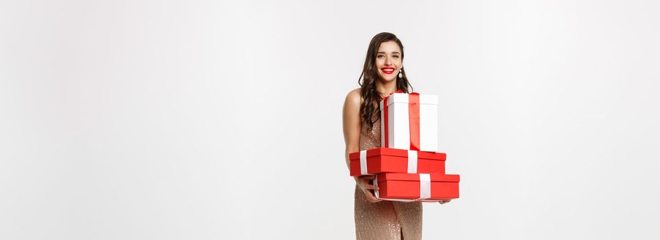 Christmas party and celebration concept. Full-length of stylish woman with red lips, glamour dress, holding gifts and smiling happy, standing over white background.