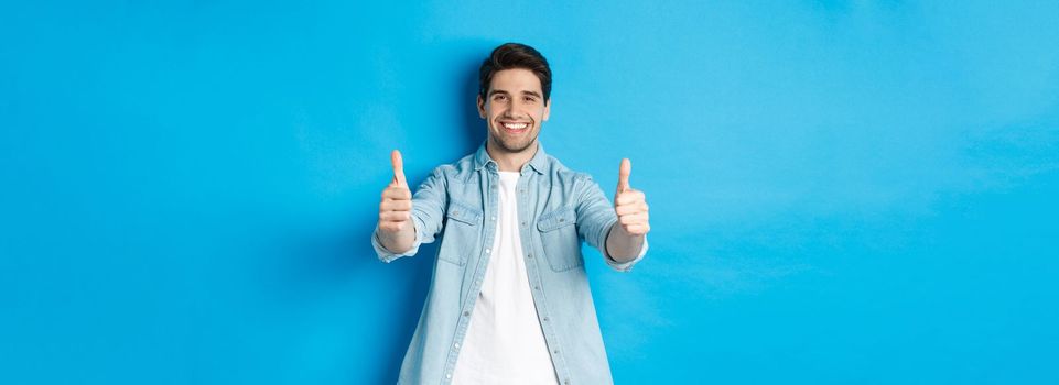 Smiling confident man showing thumbs up, guarantee quality, approving something good, standing against blue background.