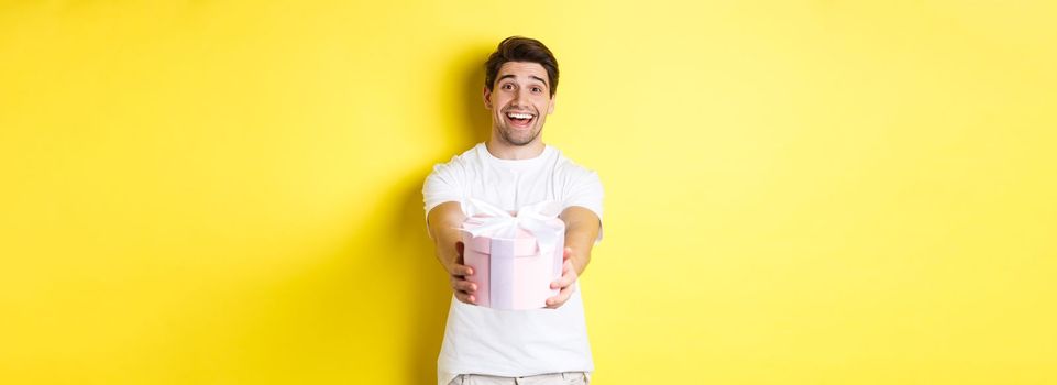 Concept of holidays and celebration. Smiling man giving you gift, congratulating, standing over yellow background with present.