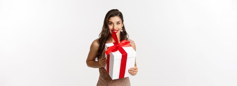 Holidays, celebration concept. Image of beautiful woman in glamour dress receive Christmas gift, looking surprised and happy, standing over white background.