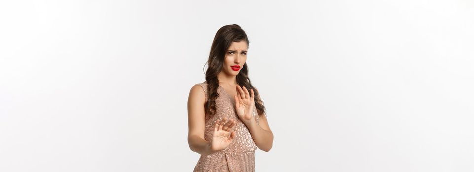 Concept of celebration, holidays and party. Woman looking displeased and rejecting something, wearing glamour dress, showing stop gesture, asking stay away, white background.