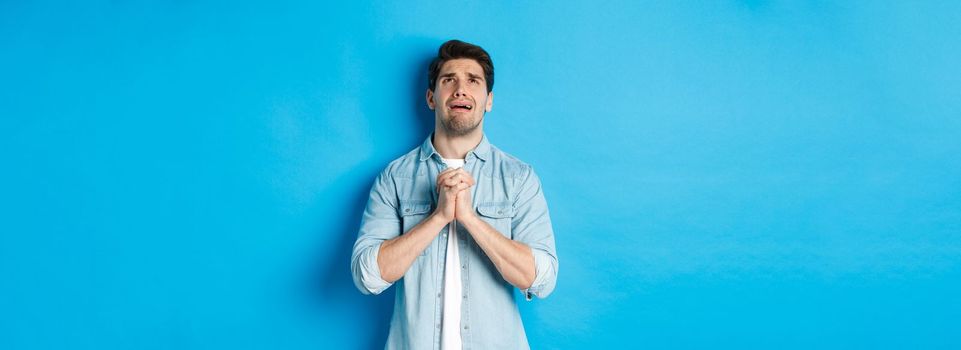 Desperate man begging and crying for help, pleading to god, looking up and holding hands in pray, standing against blue background.