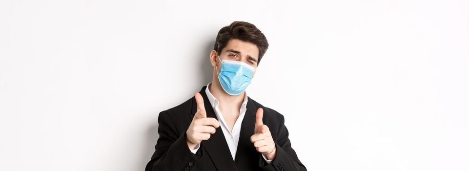 Concept of covid-19, business and social distancing. Close-up of sassy man in trendy suit and medical mask, pointing fingers at camera, standing against white background.
