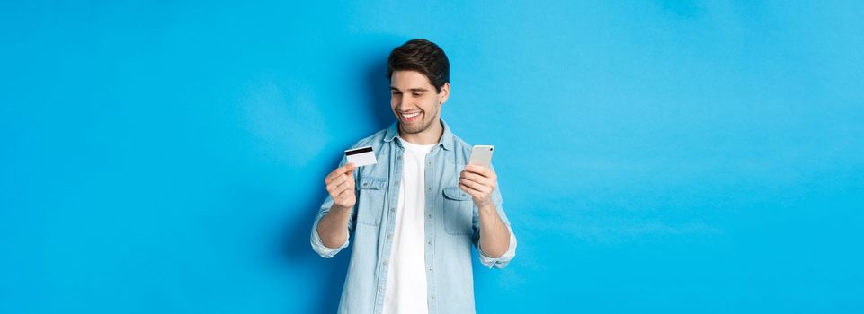 Handsome happy man paying for something online, holding credit card and mobile phone, purchase in internet, standing over blue background.