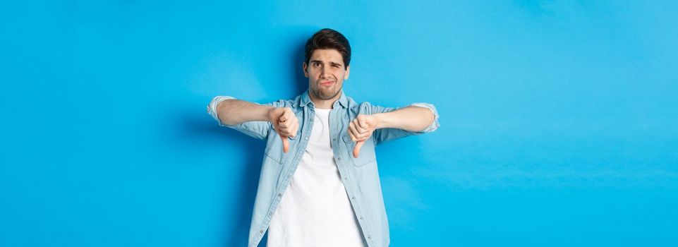 Skeptical and disappointed 25 years old man in casual outfit dislike something bad, showing thumbs-down and grimacing displeased, standing against blue background.