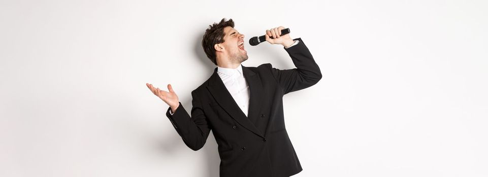Portrait of handsome man singing a song with passion, standing in black suit, holding microphone and performning, posing over white background.