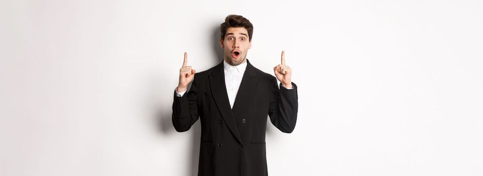 Portrait of surprised handsome businessman in black suit, saying wow and looking amazed, pointing fingers up at copy space, standing over white background.