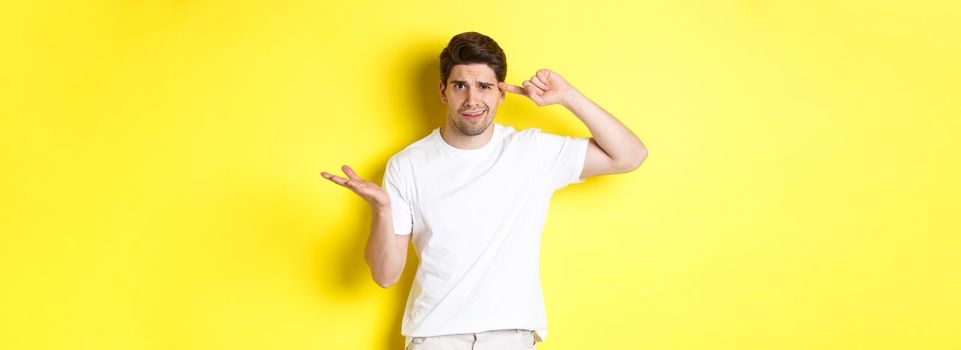 Confused and angry man pointing at head, scolding person for acting stupid, show crazy sign, standing over yellow background.