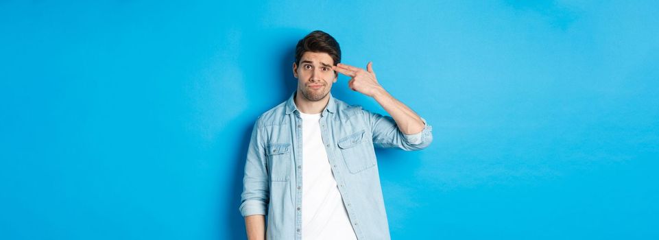 Tired and annoyed adult man making finger gun gesture near head, shooting himself from boredom, standing against blue background.