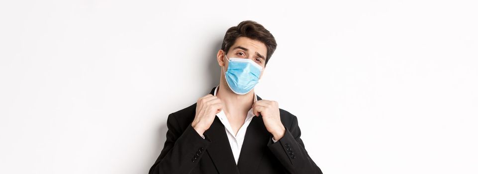 Concept of covid-19, business and social distancing. Image of confident handsome man in trendy suit and medical mask, looking sassy, standing against white background.