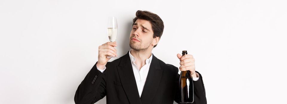 Close-up of attractive man in trendy suit, tasting champagne, looking at glass, standing against white background.