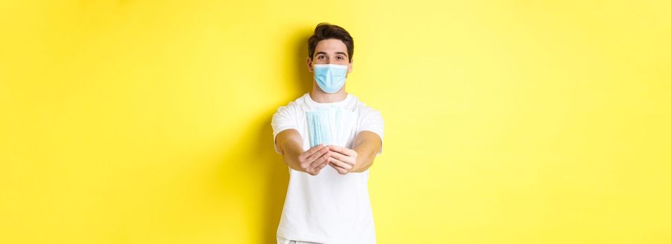 Concept of covid-19, quarantine and preventive measures. Young caucasian man giving medical masks for you, standing against yellow background.