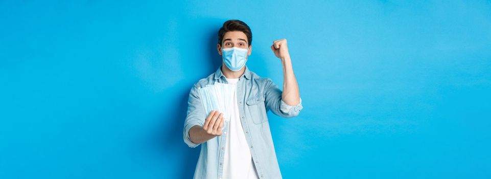 Concept of coronavirus, quarantine and social distancing. Cheerful man showing medical masks and making fist pump, celebrating or triumphing, standing against blue background.