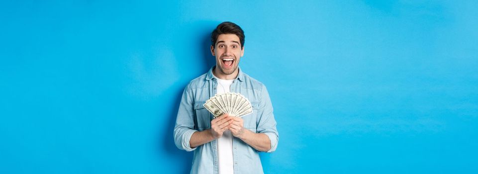 Excited and surprised attractive man, holding money prize and smiling amazed, standing over blue background. Copy space