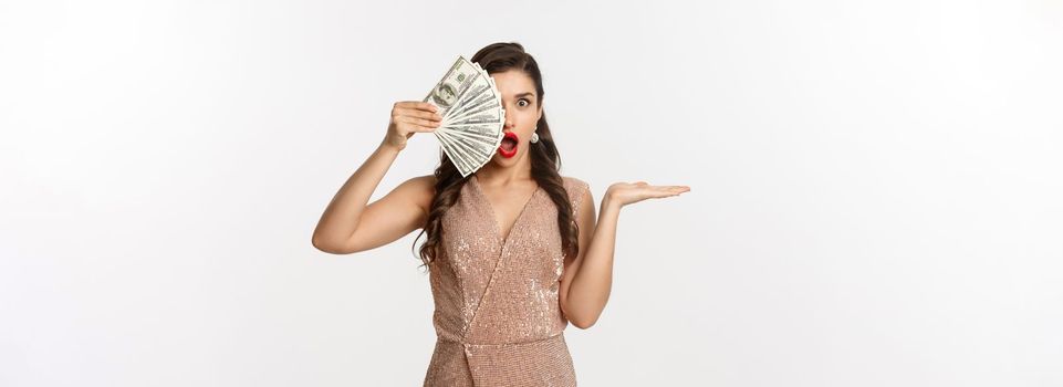 Shopping concept. Shocked woman holding money near face, wearing stylish dress, showing dollars with amazed expression, standing over white background.