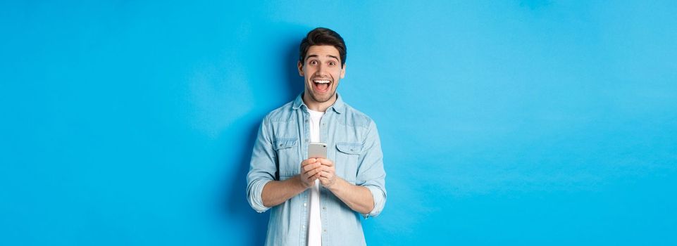 Surprised and happy man winning something online, holding smartphone and rejoicing, standing against blue background.