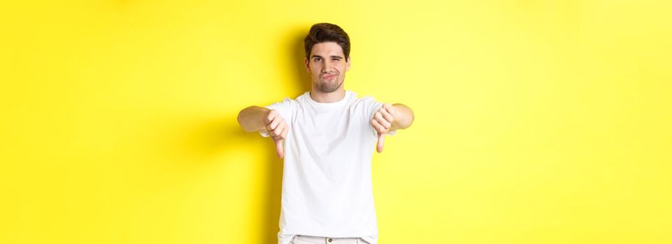 Skeptical guy showing thumbs down and smirking, dislike and disagree, standing over yellow background. Copy space