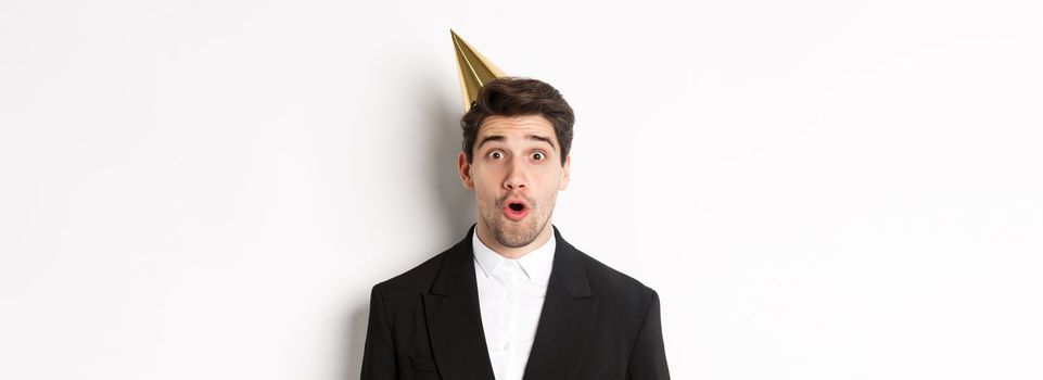 Close-up of attractive man in party hat and trendy suit, looking surprised, celebrating new year, standing against white background.