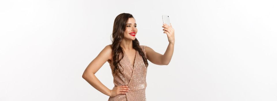 Beautiful woman with makeup and hairstyle, wearing glamour dress, taking selfie on smartphone, celebrating christmas party, white background.