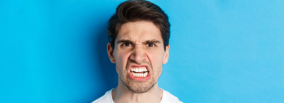 Headshot of angry caucasian man looking with scorn and dismay, being mad at person, standing against blue background.