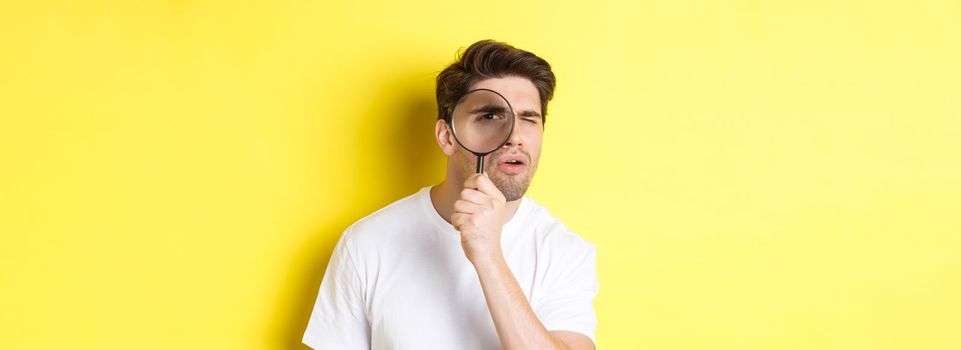 Man looking through magnifying glass with serious and thoughtful look, searching or investigating, standing over yellow background.