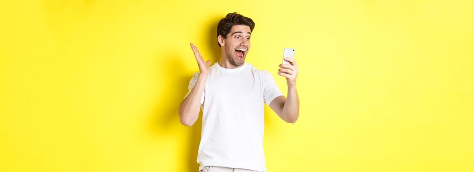 Surprised and happy man looking at mobile phone screen, reading fantastic news, standing over yellow background.