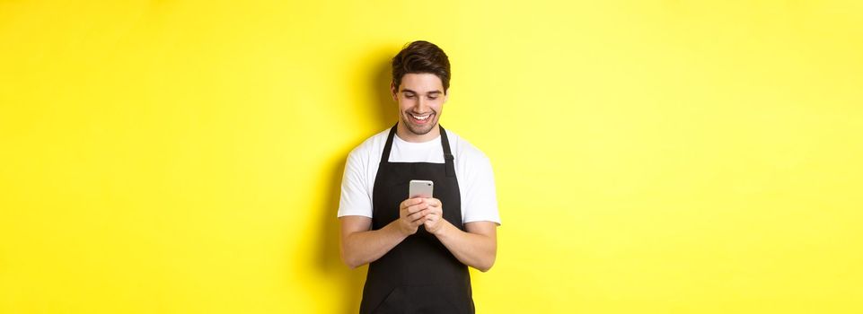 Waiter in black apron reading message on mobile phone, smiling happy, standing over yellow background.