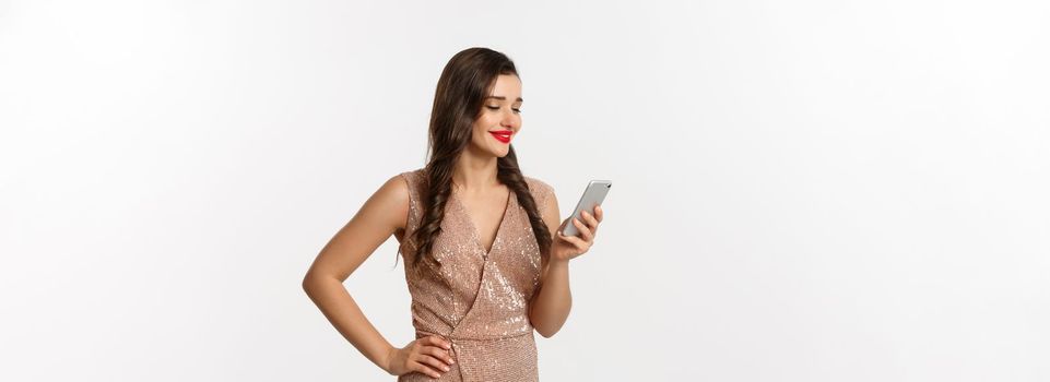 Christmas party and celebration concept. Happy young woman reading message on phone and smiling, using smartphone, wearing luxurious dress, white background.