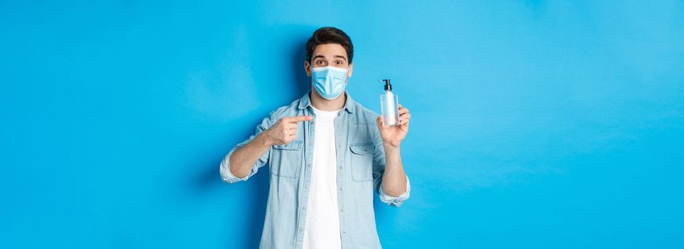 Concept of covid-19, pandemic and social distancing. Handsome guy in medical mask advice to use hand sanitizer, pointing at antiseptic, standing over blue background.