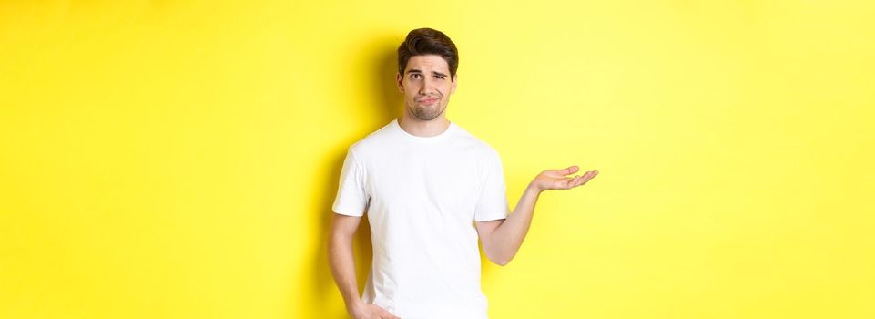 Confused and displeased guy raise hand, grimacing puzzled, standing against yellow background. Copy space