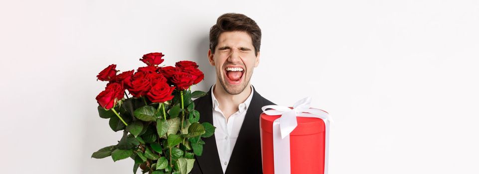 Image of sad guy in suit, got rejected and crying, holding bouquet of roses and present, standing miserable against white background.