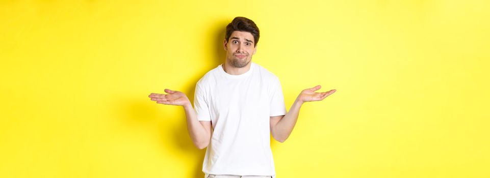 Clueless man in white t-shirt, shrugging and looking puzzled, dont know anything, standing over yellow background.