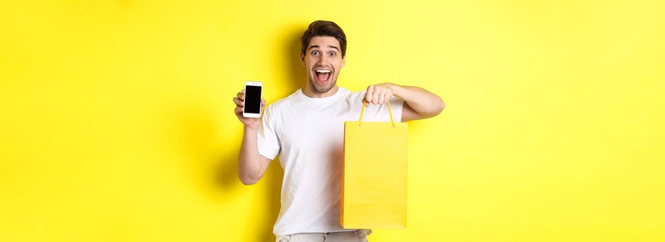 Concept of mobile banking and cashback. Young happy guy holding shopping bag and showing smartphone screen, yellow background.