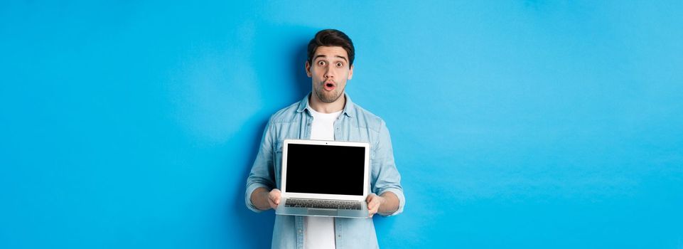 Man showing advertisement on laptop screen and looking amazed, saying wow and looking at camera, standing against blue background.