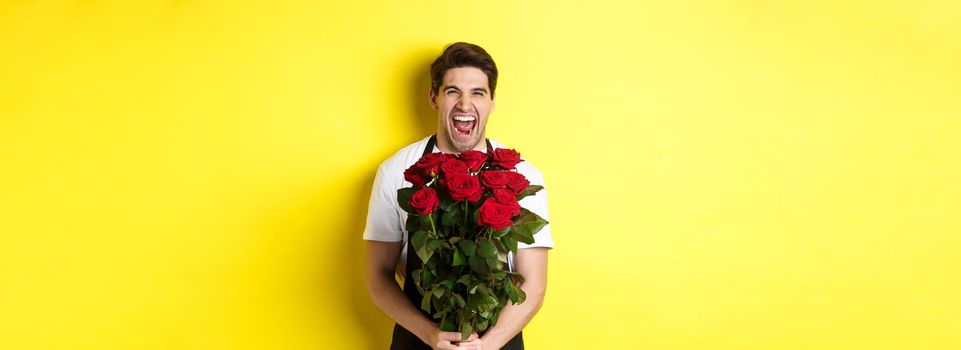 Funny young salesman in black apron holding bouquet of roses, florist laughing and standing over yellow background.