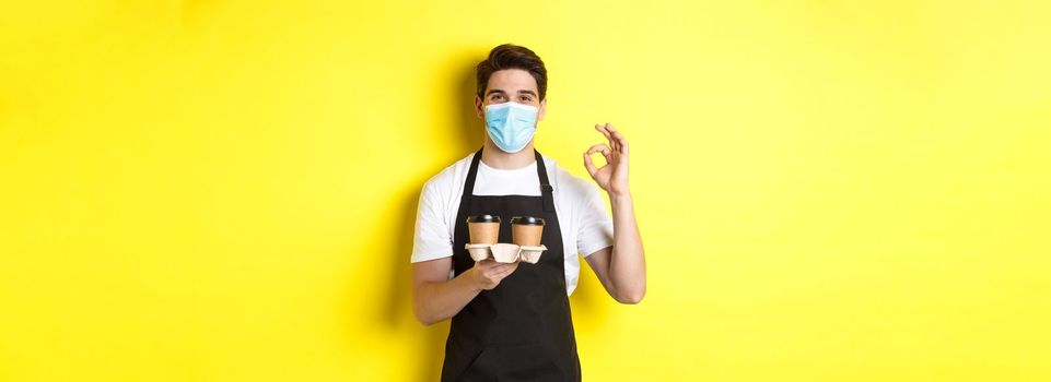 Concept of covid-19, cafe and social distancing. Barista in medical mask and black apron guarantee safety, holding takeaway cups of coffee and showing OK sign, yellow background.