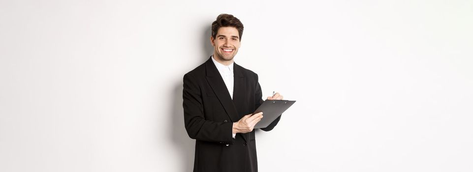 Portrait of confident businessman in black suit, signing documents and smiling, standing happy against white background.