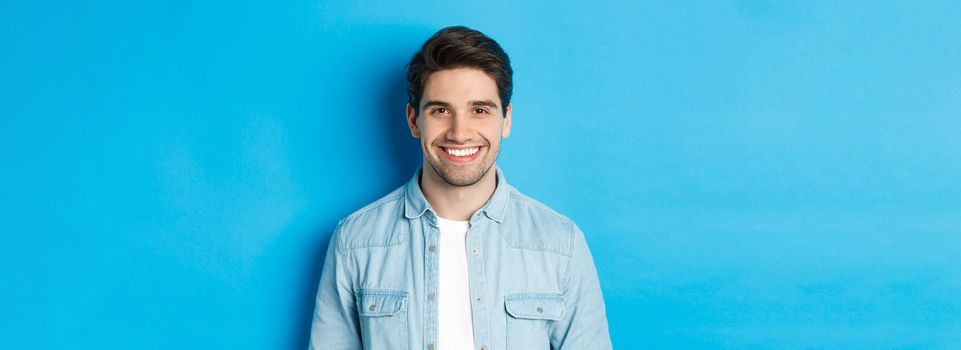 Close-up of young successful man smiling at camera, standing in casual outfit against blue background.