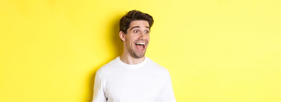 Image of happy man checking out promo, looking left with amazement, standing in white t-shirt against yellow background.