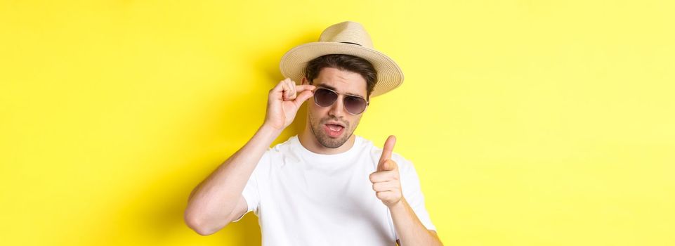 Concept of tourism and vacation. Cool and sassy man flirting with you, wearing sunglasses and pointing finger at camera, standing over yellow background.