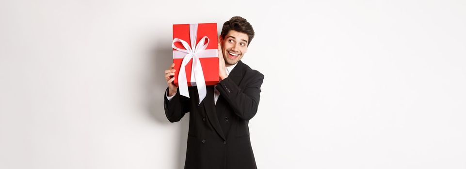 Concept of christmas holidays, celebration and lifestyle. Image of excited man enjoying new year, shaking gift box to guess what inside, standing against white background.