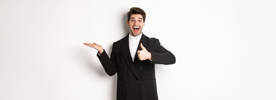 Portrait of hadnsome bearded man in formal suit, showing thumb-up and holding product in hand over white copy space, recommending product, standing over white background.