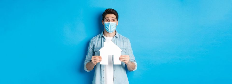 Concept of coronavirus, quarantine and social distancing. Young man searching apartment, showing house paper model, wearing medical mask, renting or buying propery, blue background.