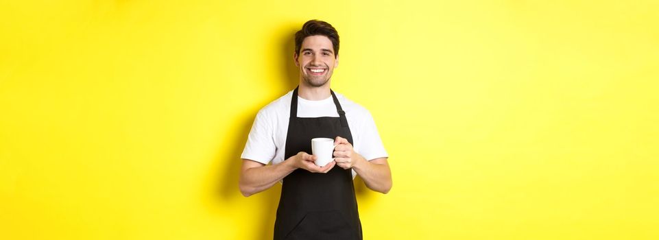 Handsome barista serving coffee, bring cup, standing in black apron with friendly smile.