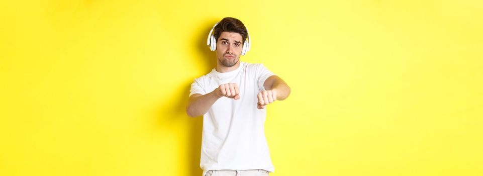 Cool guy listening music in headphones and dancing, standing in white clothes against yellow studio background.