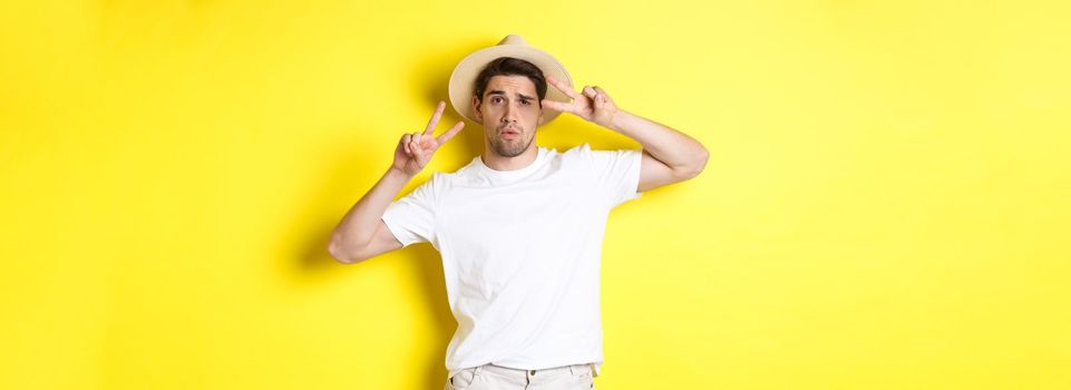 Concept of tourism and vacation. Cool guy taking photo on holidays, posing with peace signs and wearing straw hat, standing against yellow background.