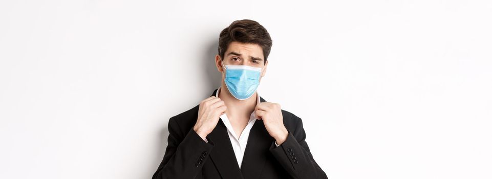 Concept of covid-19, business and social distancing. Image of confident handsome man in trendy suit and medical mask, looking sassy, standing against white background.